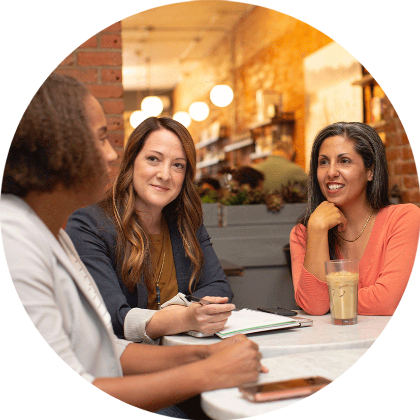 Three women sit at a cafe table, engaged in conversation. One woman holds a pen and notebook, while another has a glass of iced coffee. The setting is cozy, with warm lighting and a blurred background.